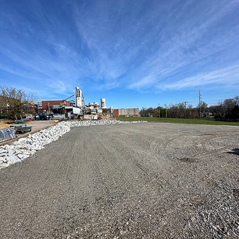Industrial construction site with a gravel lot, rock piles along a low wall, and a factory building under a blue sky.